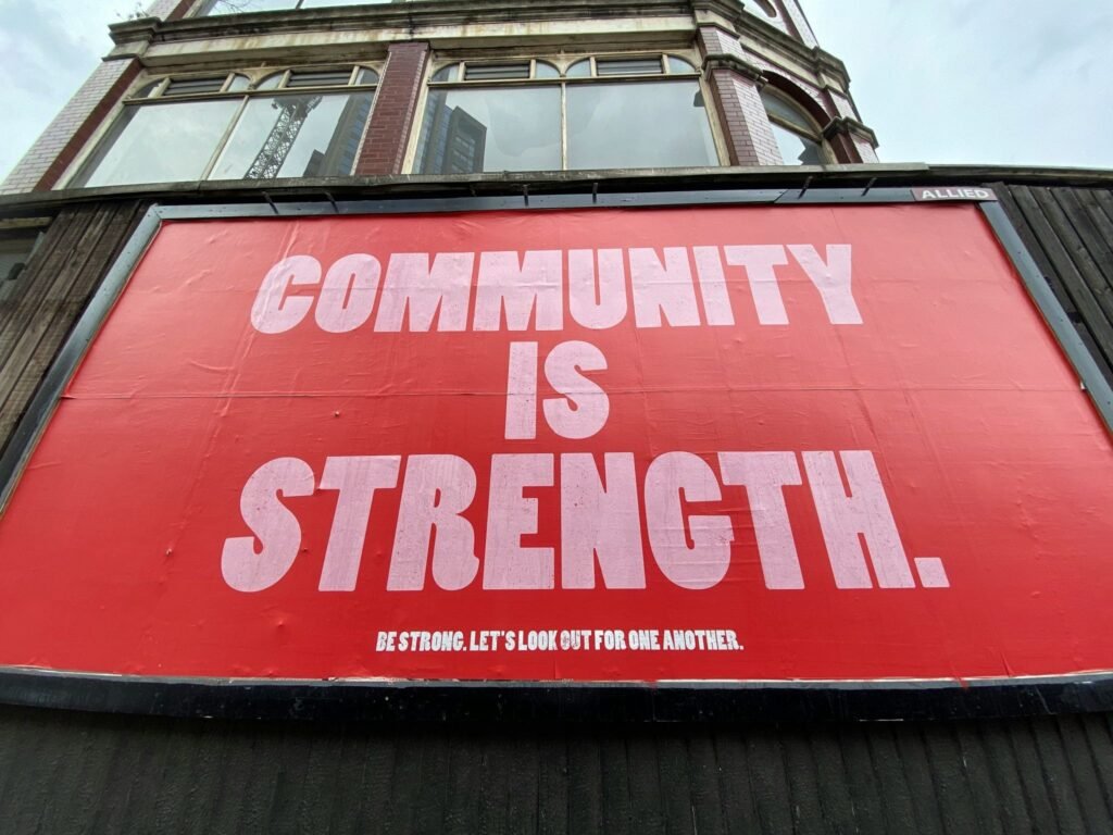A large red sign mounted on the side of a building reads "Community Is Strength" in large white letters, with a smaller message below encouraging people to look out for one another.